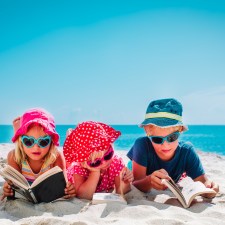 Three young children reading books on a beach. Books for your kids' summer reading list include stories about animals, school and more.